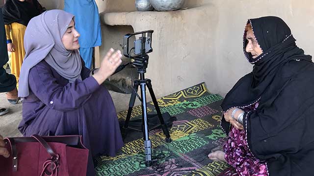 A VOA journalist uses her cell phone mounted on a tripod to interview an older woman seated on the floor.