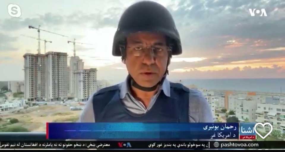 A screenshot of a VOA journalist wearing a helmet and bullet-proof vest reports from in front of an Israeli skyline at sunset.
