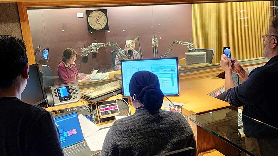 People inside of a recording studio, watch two VOA journalists recording a show.