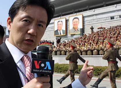 A reporter holds a VOA microphone while covering a military parade with North Korean soldiers marching down a street.