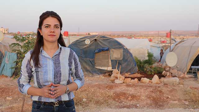 Photo of a journalist standing in front of a refugee camp.
