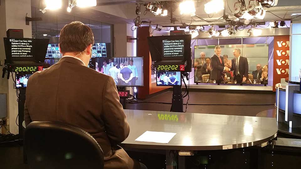 Photograph of the back of VOA journalist broadcasting from inside of a studio with television screens and teleprompters.