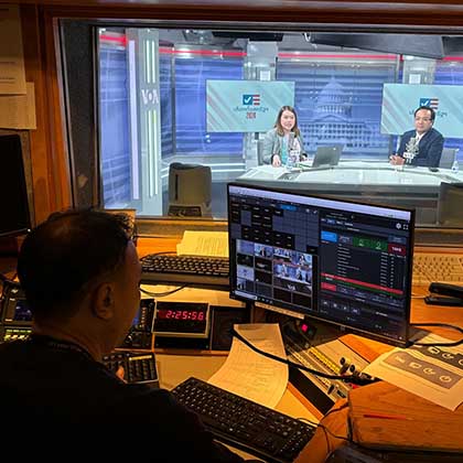 Photo of a man inside the broadcast control room watching two journalists in the studio.